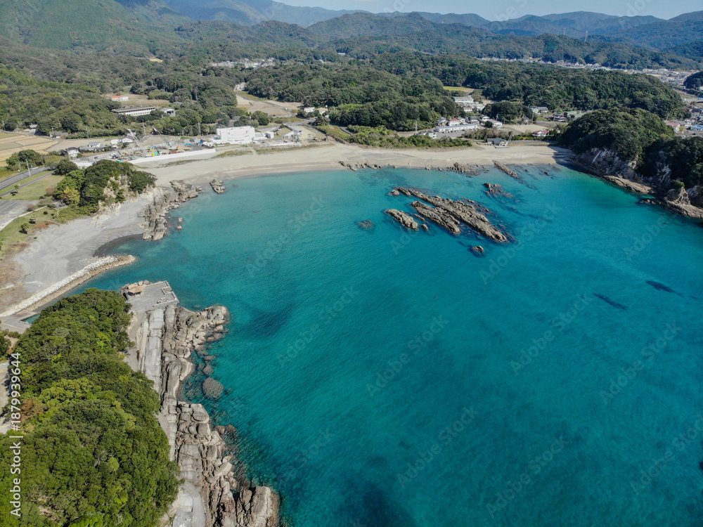 Obraz premium Wide aerial view of a turquoise coastal bay with rocky formations, sandy beach, and forested hills along the Shikoku coastline in Japan