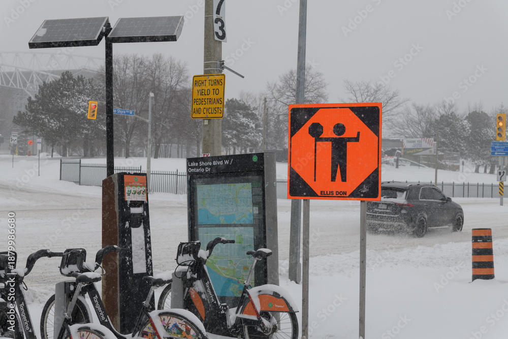 Fototapeta premium orange construction sign, Bike Share Toronto docking station and map at intersection of Lake Shore Blvd W and Ontario Drive during a snow storm on 2026-01-15