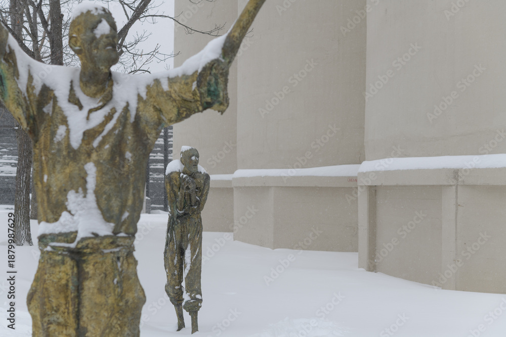 Fototapeta premium Apprensive Man, with defocused The Jubilant Man by Rowan Gillespie part of The Arrival, set beside the old Canada Malting silos on Eireann Quay, Toronto, during winter storm on 2026-01-15