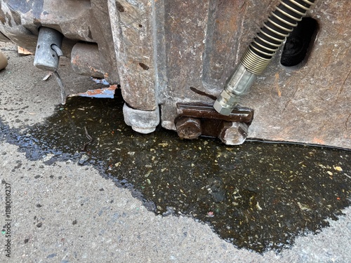 Close-up of leaking hydraulic oil beneath heavy construction machinery, showing metal fittings, hoses, and an oil spill spreading across concrete ground.
