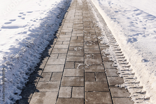 Snow cleared sidewalk with paving stones in winter. Pedestrian path after snowfall with snowbanks on both sides. Urban walkway safety concept. Cold season street maintenance and accessibility