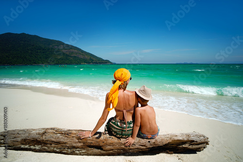 Rear view of a mother and child sitting together on a sandy tropical beach and looking at the turquoise sea. Summer vacation, family travel and lifestyle concept. 