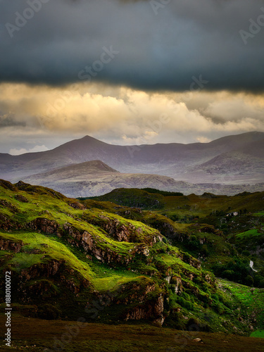 A green, vegetated hillside with rocky outcrops slopes down into a valley. In the background, distant mountains rise under a cloudy sky in the national park.