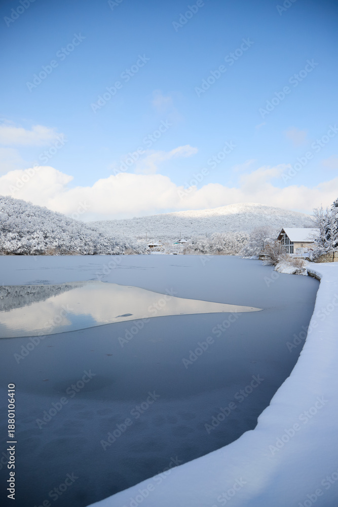 Obraz premium A calm winter landscape on the lake in the village of Semigorsky in the mountains of the city of Novorossiysk, snow-covered shores, the silence of nature and a cold atmosphere.