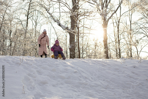 Young mother and little daughter with yellow sled standing on top of snowy hill in winter park