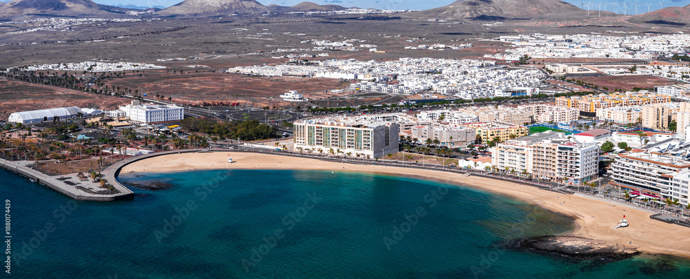 Fototapeta premium Aerial view of Arrecife, Lanzarote, shows Playa del Reducto, turquoise water, promenade, mid rise hotels, white buildings, volcanic hills, wind turbines, bright daylight.