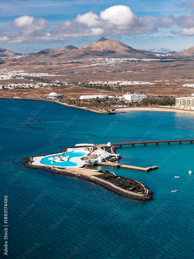 Fototapeta premium Aerial view of Islote de Fermina saltwater pools in Arrecife, Lanzarote, with white architecture, palm terraces, a long pier, marina, and volcanic peaks in midday light.