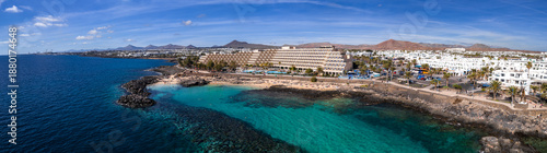 Schilderij op canvas Aerial view of Puerto del Carmen in Lanzarote shows a volcanic shore, a sandy cove, a terraced beachfront hotel, palm lined promenade, and rust red cones near Arrecife