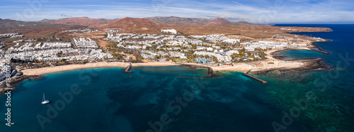 Foto Aerial view of Costa Teguise, Lanzarote, shows white buildings, golden coves, turquoise shallows, curved breakwaters, a marina with a lone catamaran, and ochre volcanic hills