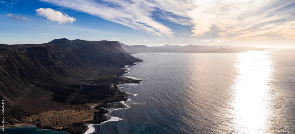 Fototapeta premium Aerial view shows Famara cliffs on Lanzarote, the strait to La Graciosa, dark lava fields, black rock shore, distant volcanic cones, and wispy high clouds at late afternoon.