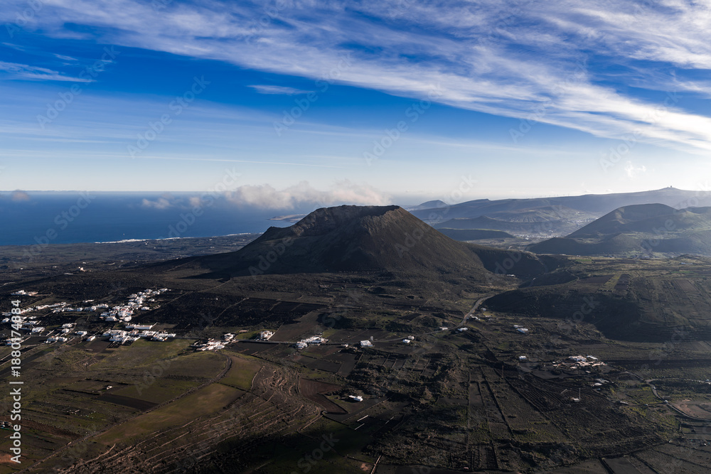 Obraz premium Aerial view of Lanzarote, Canary Islands, with a cinder cone, patterned lava, patchwork farmland, whitewashed villages, and the Atlantic along the left horizon.