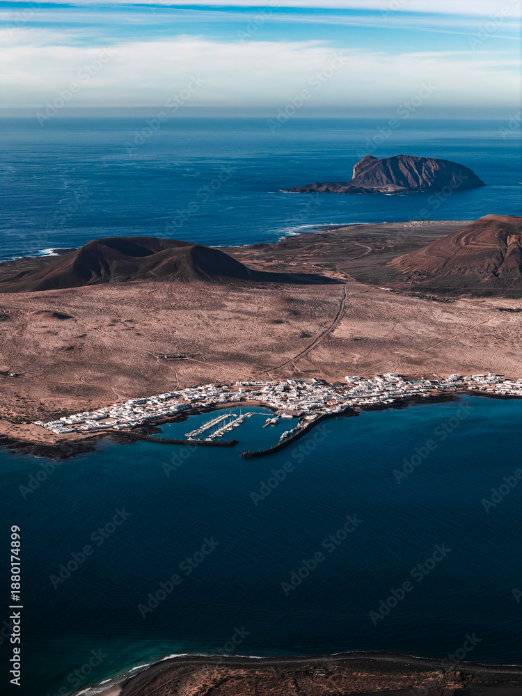 Fototapeta premium Aerial view of a whitewashed village and marina on north Lanzarote, Canary Islands, with cinder cones, lava fields, turquoise shallows, and La Graciosa on the horizon.