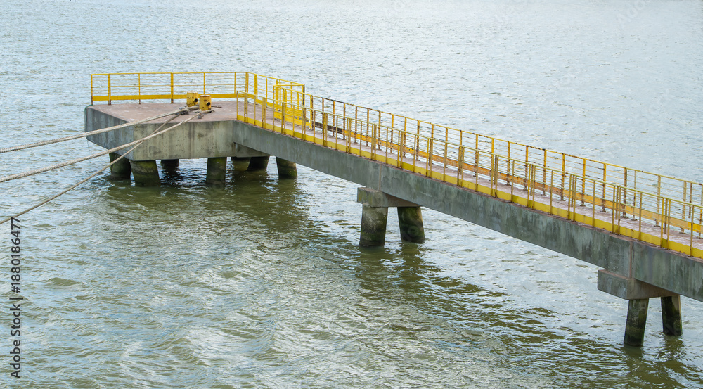 Obraz premium Concrete industrial pier with yellow railings on murky water. Mooring ropes attached to bollards. Minimalist maritime infrastructure for shipping and transport