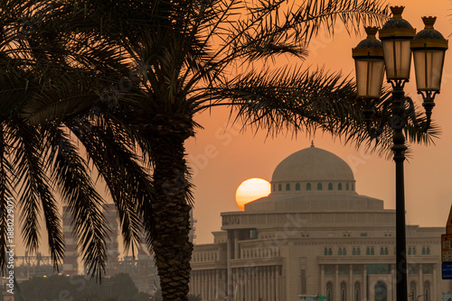 A sunset occurs behind a government building. Palm trees frame the scene. Street lamps are visible as the sun sets on the horizon.