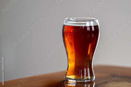 A single beer sample on a wooden table with a grey background. The alcoholic beverage is in a clear pilsner glass. The amber color sampler is a carbonated, malty beer. The glass is part of a flight.