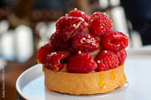 A single round glazed raspberry almond tart on a white dessert plate is being served on a terrace of a fancy patisserie. The tartlet has whole bright red raspberries and a crust filled with cream. 