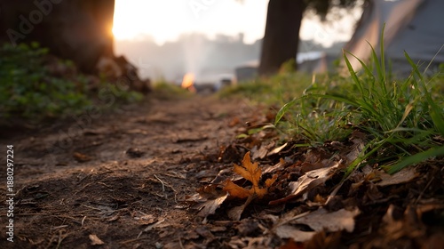 A dirt path covered in autumn leaves leads towards a serene campsite with a gentle sunset glow and faint smoke in the distance