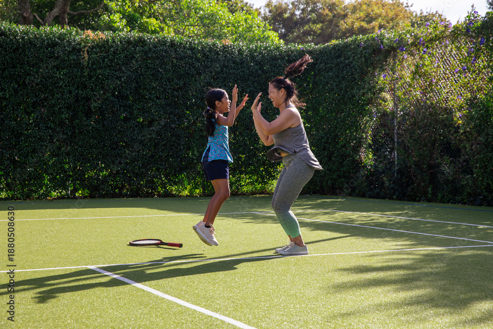 Obraz premium Diverse mom and daughter in gear jumping and high-fiving on tennis court with racket, white lines