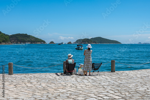Wallpaper Mural Under a clear blue sky at Repulse Bay, two people relax on a stone promenade with fishing gear. Vibrant blue sea dotted with boats and framed by lush green islands in the distance. Hong Kong Torontodigital.ca