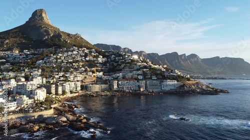 Cinematic drone footage of the picturesque Camps Bay coastline and Victoria Road winding between the ocean and mountains; aerial shot of the famous white sand beach and luxury apartments in Cape Town