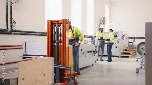 Male factory worker in safety jacket and helmet operating orange pallet lifter near machinery. In background colleagues in discussion using laptop. Industrial workspace with equipment and materials.