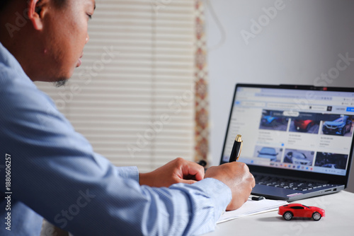 Buying and selling cars online. A man wants to buy a used car. He browses various online used car listings on his laptop to compare prices. He then notes it down.