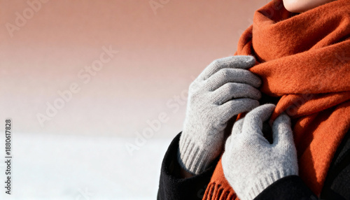Woman wearing grey knitted gloves adjusts a warm orange wool scarf. Person bundling up in cozy winter fashion outdoors. Cold weather seasonal apparel with copy space