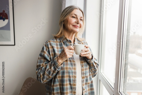 Adorable caucasian smiling senior lady in plaid shirt enjoying her herbal tea standing in cozy room next to big window and admiring first snow and winter coming looking through at city street