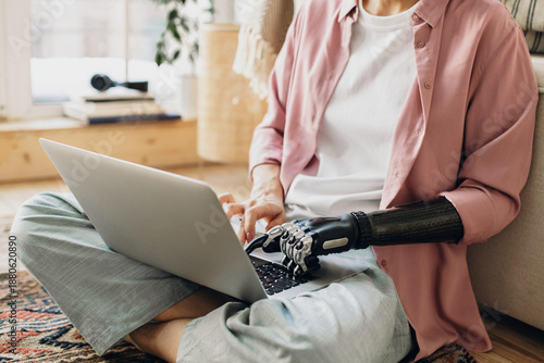 Crop shot of robotic prosthesis hand of female working on laptop in living room , sitting on floor against window, dressed in casual clothes. Cybernetics, high technologies, bio-engineering