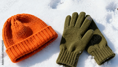 A vibrant orange knit beanie and a pair of green gloves rest on white snow. Essential winter accessories for cold weather. Warm clothing flat lay