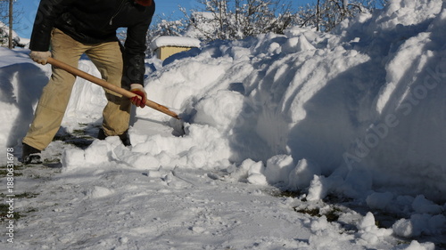 A man, visible without a face, clears large snowdrifts in front of a house with a wooden shovel in sunny weather. Manual snow removal from a house's surrounding area after a storm.