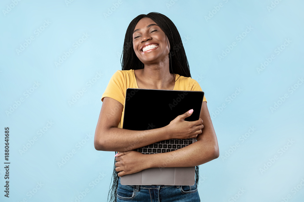 Obraz premium People And Gadgets. Joyful Black Woman Hugging Laptop Standing In Studio Over Blue Background. Lady Posing Holding Computer. Freelance And E-Learning. I Love Internet Technologies Concept