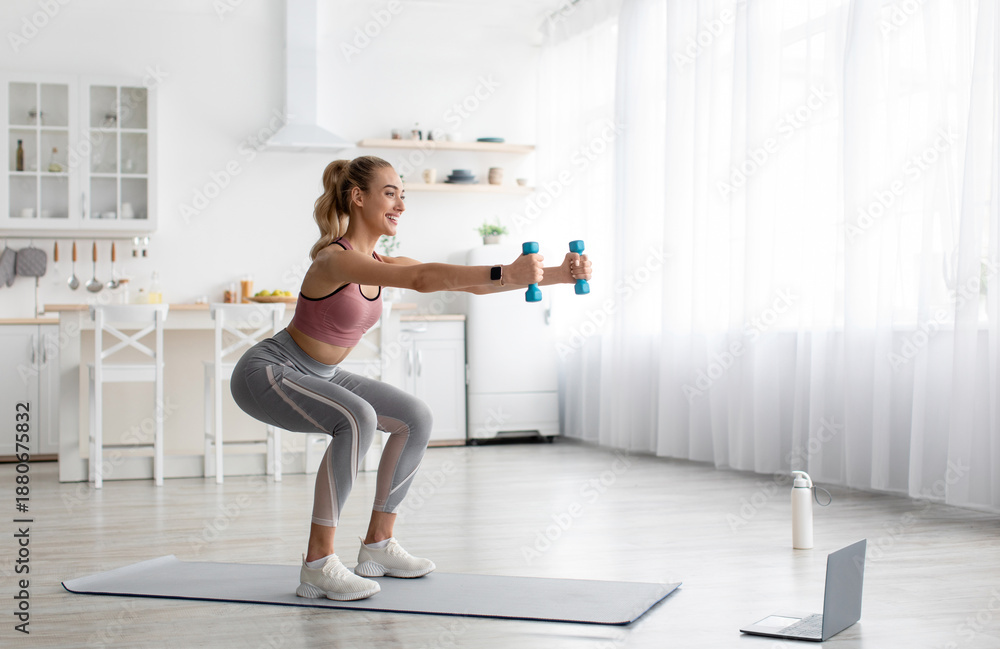 Obraz premium Keep fit, fitness or pilates with online class, morning workout at home. Happy young blonde female in sportswear in minimalist living room interior doing squats with dumbbells on mat with laptop