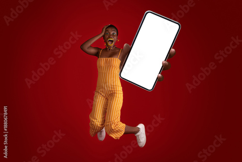 Emotional stylish millennial african american woman jumping up with modern cellphone with blank screen, short-haired lady with bright makeup recommending newest mobile app, red background, mockup