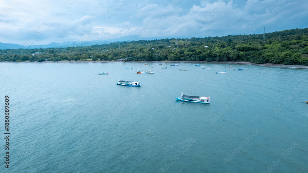 Fototapeta premium High angle landscape of various fishing boats floating on tranquil turquoise sea near a lush green coastline under a cloudy sky in summer morning. 