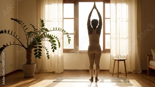 Yoga practitioner standing on mat facing sunlit window, al fresco calm interior with plant and minimal furniture