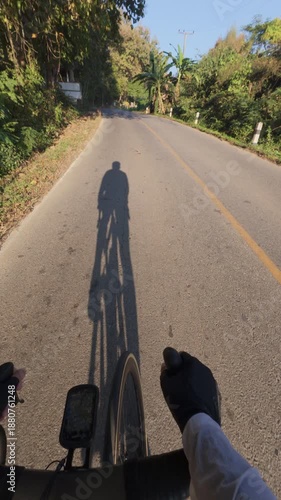 A cyclist's shadow on a sunlit road, surrounded by lush greenery and trees, capturing the essence of outdoor adventure.
