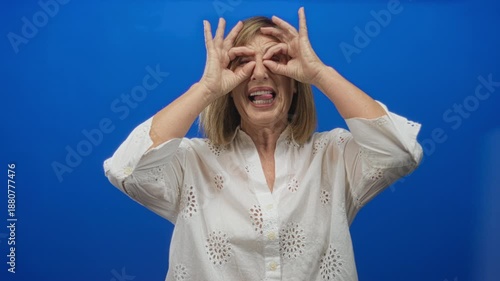 Caucasian middle aged woman in white blouse smiling frames eyes with hand binoculars gesture in studio; playfulness.