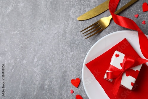 Top view of cutlery with a napkin, plate, gift box and decorative hearts on a gray table with space for text. Valentine's Day romantic dinner