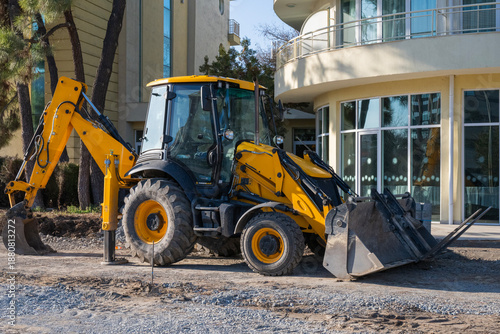 Fotografía a yellow backhoe loader parked on a dirt path in front of a modern beige residential building with large glass windows