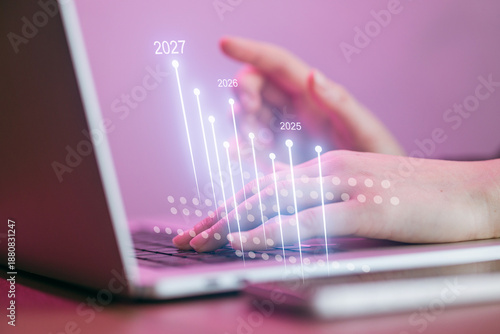 Hands typing on a laptop with digital graphs and numbers showing future years in a workspace setting
