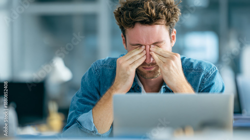 Young man wearing a denim shirt rubbing his eyes while working on a laptop in a modern office environment showing signs of fatigue and stress