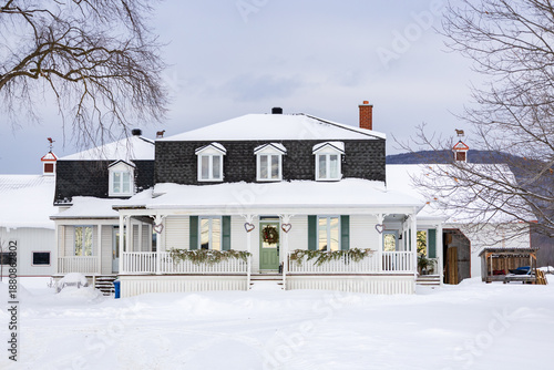 Beautiful white clapboard country house with black Mansard shingled roof surrounded by farm buildings seen during a cloudy winter morning, Quebec City, Quebec, Canada