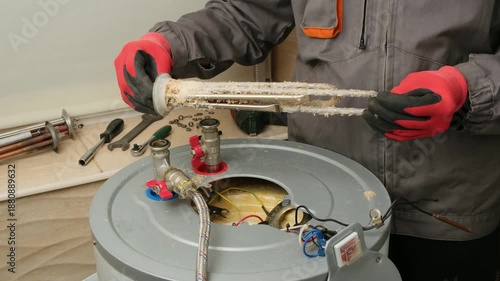 Repairman in red gloves holding and rotating a burnt-out boiler heating element covered with lime coating. Electric Water Heater Maintenance and Repair.