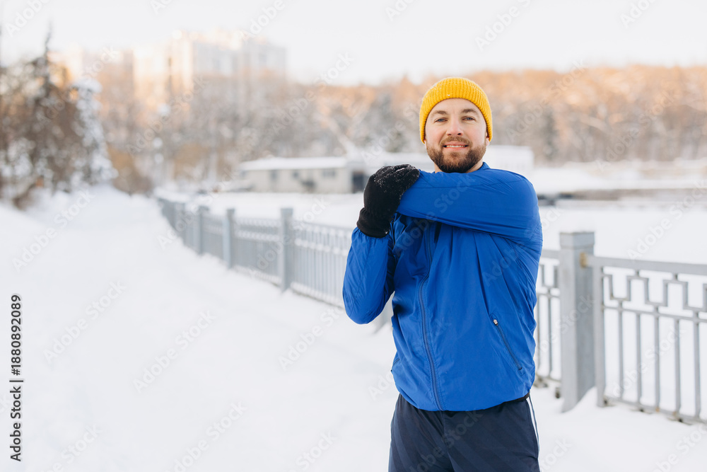 Naklejka premium Man stretching arm in winter park, preparing for outdoor running or fitness exercise in snow