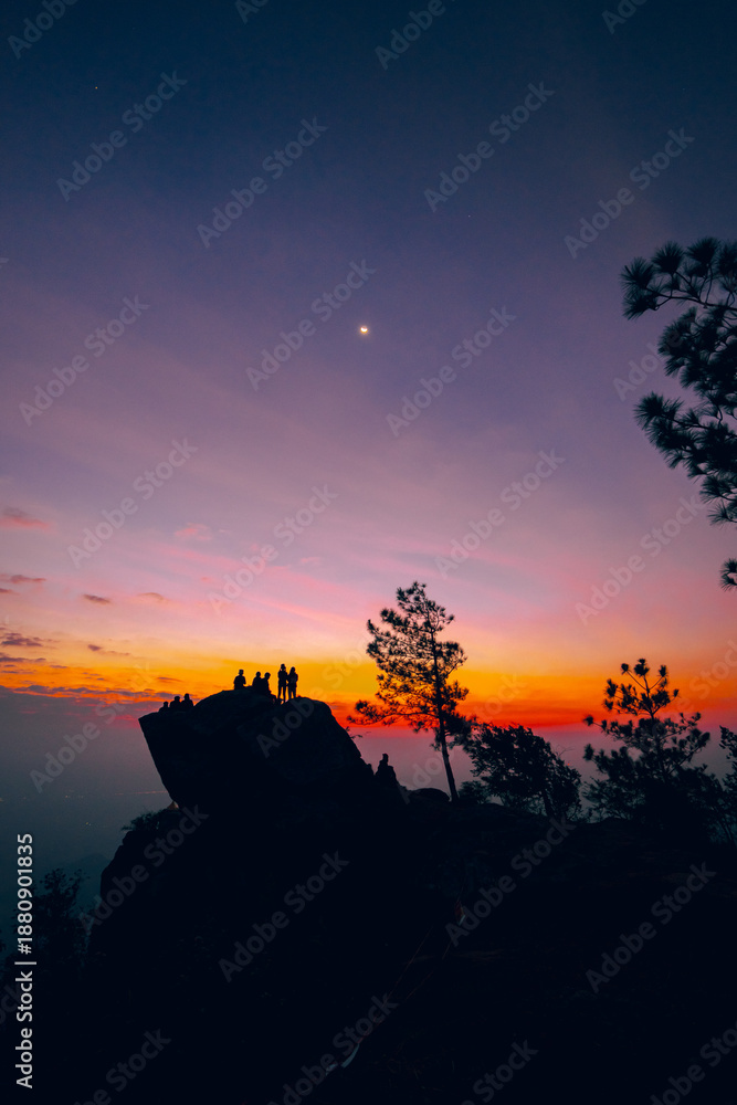 Obraz premium Silhouette of a group of people climbing on a large cliff. Colorful sky of orange, yellow, pink, and blue in the morning at sunrise. There are mountains and trees around with copy space on top.