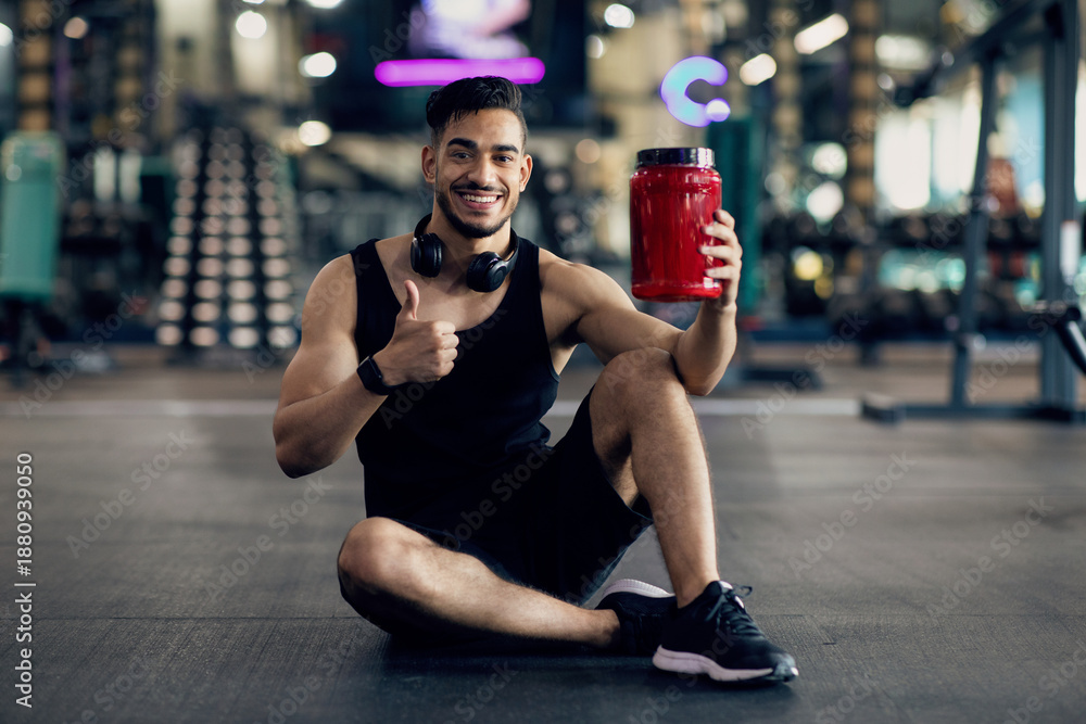 Fototapeta premium Handsome Muscular Middle Eastern Guy Advertising Fitness Supplements While Posing At Gym, Young Arab Male Athlete Holding Red Container With Whey Protein Powder And Showing Thumb Up At Camera