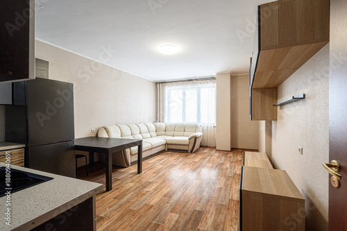 compact living space with a beige sectional sofa, dark wooden table, built-in wooden shelves, light wood flooring, large window with curtains, and a modern kitchen area