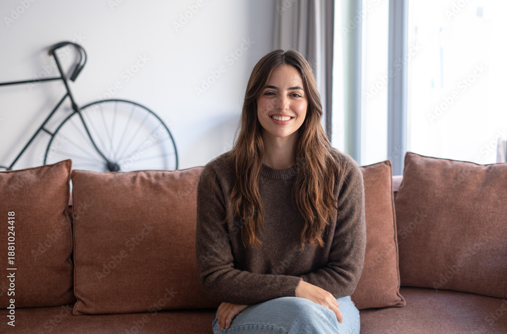 © luciano - Portrait of a smiling young woman with long brown hair sitting on sofa in a cozy modern living room looking at camera. Natural light, warm tones. Concept of happiness, confidence, and everyday life © luciano - Portrait of a smiling young woman with long brown hair sitting on sofa in a cozy modern living room looking at camera. Natural light, warm tones. Concept of happiness, confidence, and everyday life