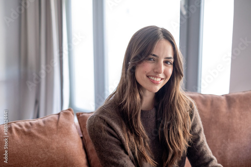 фотографія Portrait of a smiling young woman with long brown hair sitting on sofa in a cozy modern living room looking at camera